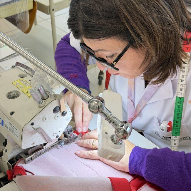 Profilo di una sarta modellista con occhiali e camice bianco concentrata alla macchina da cucire professionale mentre assembla un capo rosa.