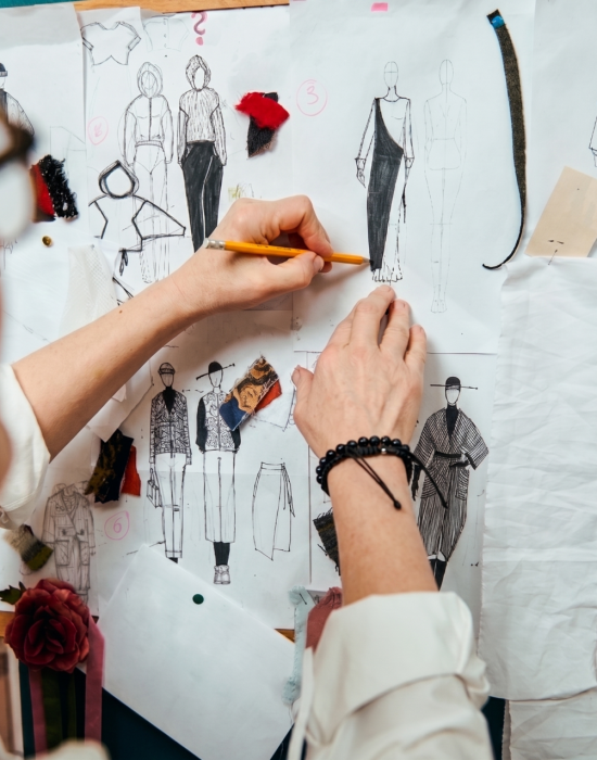 Soft focus on the fashion designer's hands holding a pencil and sketching clothes on paper pinned to the wall with drawings and esquises attached. Creative professional occupation concept